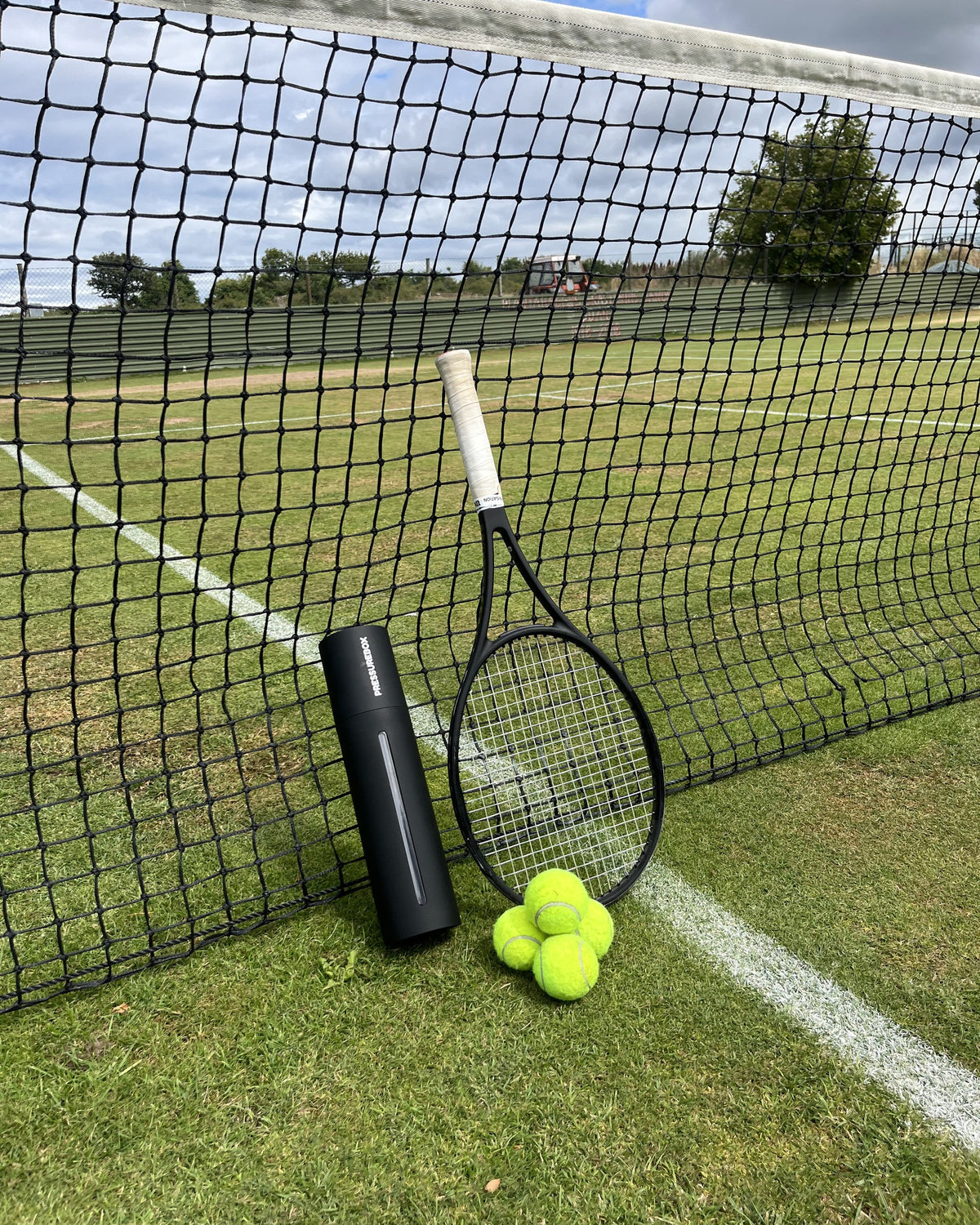 Pressurebox Pro Tennis automatic ball pressurizer leaning against the net on a grass tennis court, next to a racket and four tennis balls