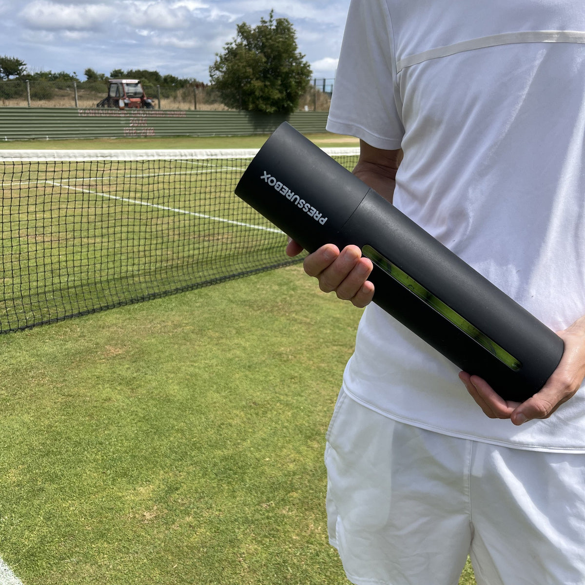 A tennis player in white holding a Pressurebox Pro Tennis ball pressurizer by the net on a grass court